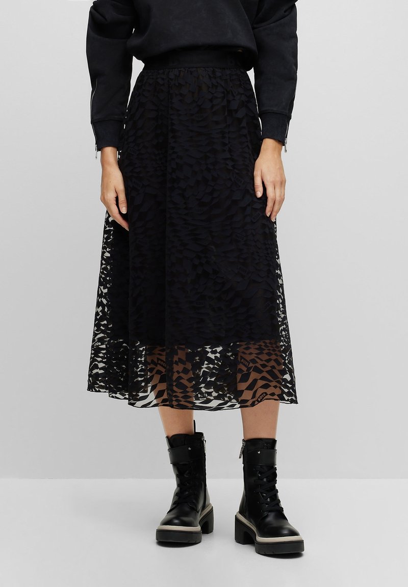 Femme portant une jupe midi noire texturée avec un surjet transparent à motifs, associée à des bottines noires épaisses à lacets.