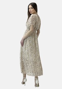 Abito maxi floreale con maniche corte, tessuto beige chiaro, dettagli dorati, vita elastica e gonna fluida, abbinato a tacchi bianchi.
