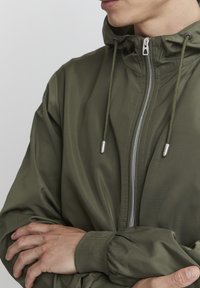 Veste légère vert olive avec fermeture éclair, capuche réglable, poignets élastiques et texture lisse. Aucun motif ni logo visible.