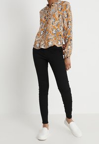 Bluzka z długim rękawem w musztardowym kolorze z kwiatowym wzorem, zestawiona z czarnymi dopasowanymi dżinsami i białymi butami wsuwanymi. Gładki materiał; luźny krój.