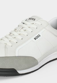 Basket blanche BOSS avec lacets blancs, panneau latéral texturé, bout en daim gris et semelle en caoutchouc noire visible sur fond blanc.