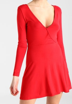 Kobieta ubrana w dopasowaną, czerwoną sukienkę z długim rękawem, głębokim dekoltem w kształcie litery V i rozkloszowanym dołem na jednolitym jasnym tle.
