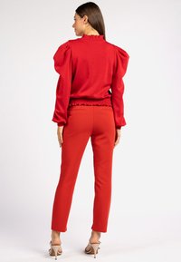 Roter Pullover mit Puffärmel und gerafftem Ausschnitt, kombiniert mit eng anliegenden roten Hosen. Das Material scheint weich zu sein; die Schuhe sind helle, hochhackige Schuhe.