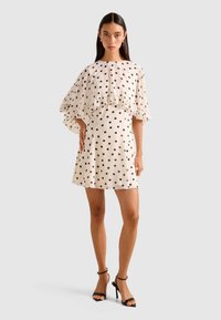 Woman wearing a cream-colored, short polka dot dress with ruffled cape sleeves, paired with black high-heeled sandals, standing on gray background.