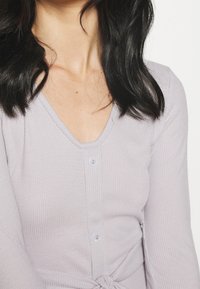 Hellviolettes, geripptes Oberteil mit V-Ausschnitt, drei Knöpfen und einer gebundenen Detail am Taillenbereich. Weiche Textur und figurbetontes Design.