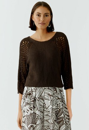 Vrouw draagt een donkerbruine gebreide trui met opengewerkte mouwen en een witte rok met grote bruine bloemmotieven, staand tegen een effen achtergrond.