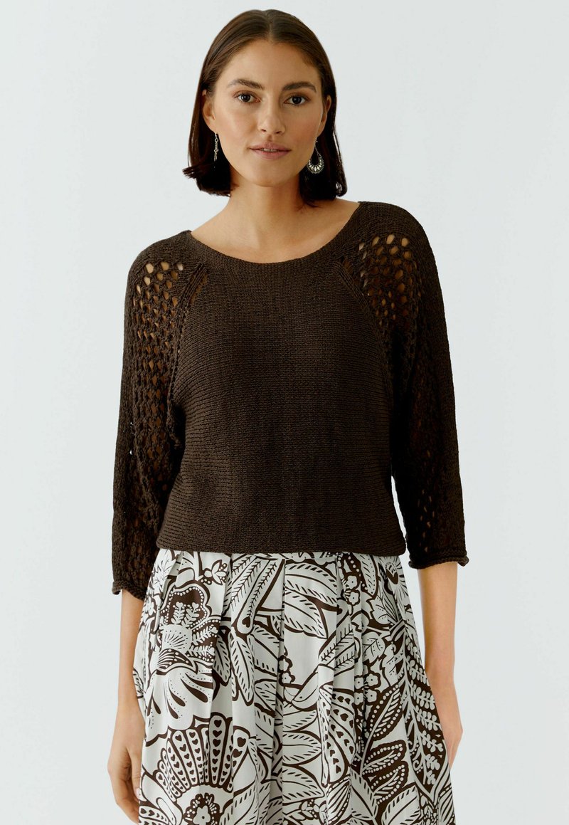 Femme portant un pull en maille marron foncé avec des manches à jours et une jupe blanche ornée de grands motifs floraux marron, se tenant devant un fond uni.