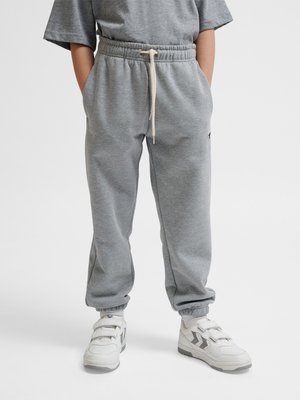 Bambino che indossa pantaloni della tuta grigi con coulisse in vita, mani nelle tasche e scarpe da ginnastica bianche con dettagli grigi, in piedi su sfondo bianco.