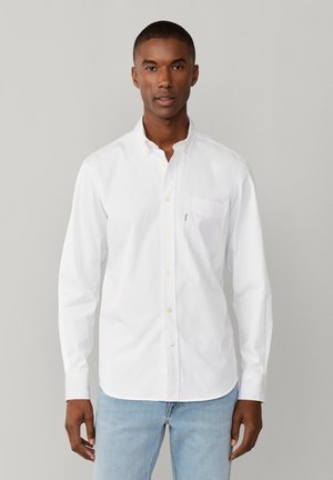 Homme portant une chemise blanche à boutons avec une poche poitrine et un jean bleu clair, debout devant un fond gris uni.
