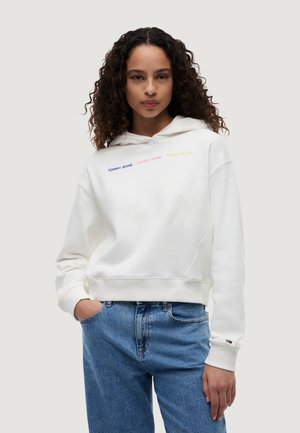 Femme aux cheveux bouclés portant un sweat à capuche blanc avec un texte multicolore "Tommy Jeans" et un jean bleu, debout avec une main dans la poche.