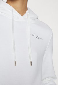 Felpa bianca con cappuccio realizzata in morbido cotone. Presenta un logo frontale in nero, lacci con punte metalliche e una superficie texturizzata.