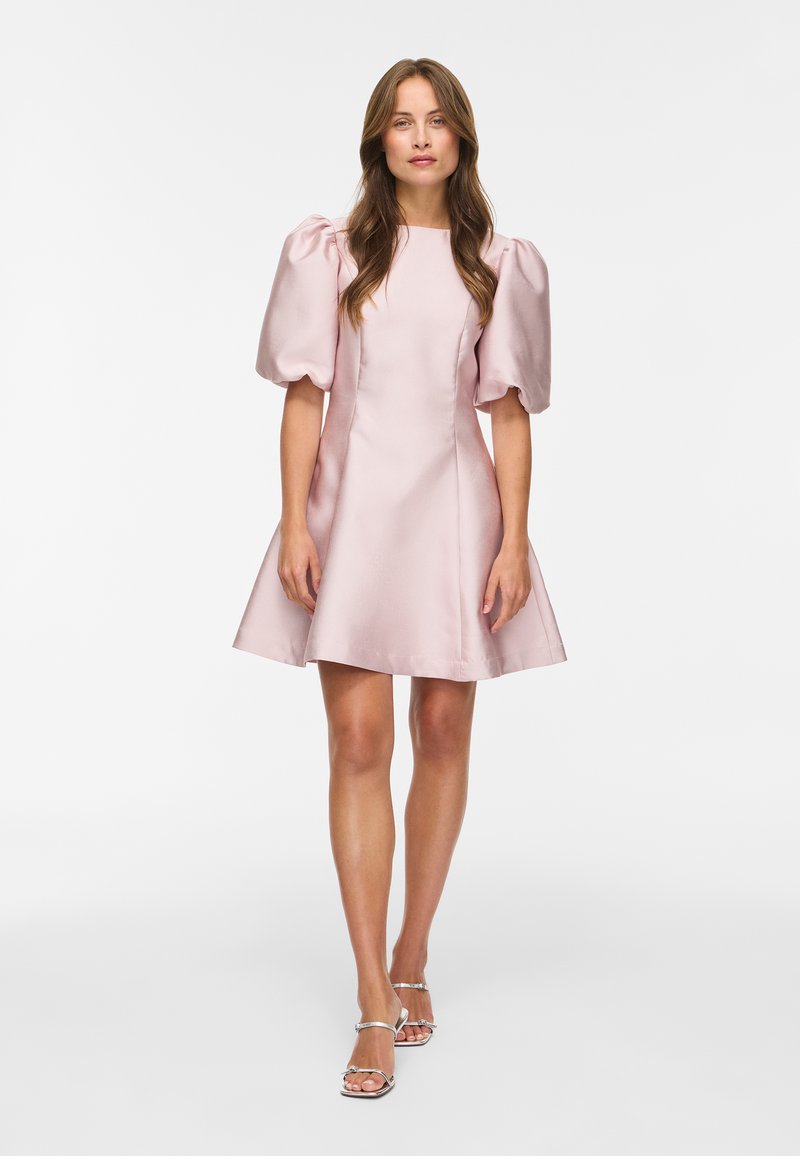 Femme debout portant une robe en satin rose clair avec des manches courtes bouffantes et des talons ouverts argentés sur fond blanc.