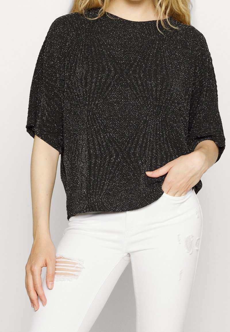Femme portant un haut noir à manches courtes pailleté avec un jean blanc déchiré, main dans la poche, sur un fond uni.