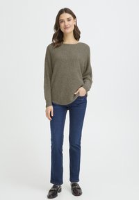 Pull texturé vert olive avec un large décolleté, associé à un jean bleu foncé et des mocassins noirs ornés de détails dorés.