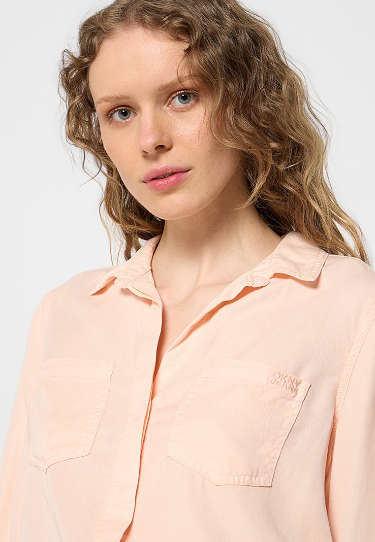 Joven mujer con cabello rubio ondulado que lleva una camisa de color durazno claro con botonadura y dos bolsillos en el pecho, uno de ellos bordado con "DKNY JEANS".
