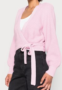 Cárdigan envolvente rosa tejido, con mangas largas, textura acanalada y cierre de lazo en la cintura. Combinado con pantalones negros de tiro alto.