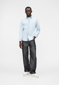 Camisa azul-clara com botões e mangas compridas, combinada com jeans de ganga escura e ténis brancos com detalhes em bege. Design simples e limpo.