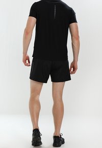 Camiseta atlética negra de manga corta con un detalle de raya vertical en la parte trasera, combinada con pantalones cortos negros y zapatillas deportivas negras.