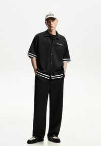Camicetta nera a maniche corte con strisce bianche e bottoni, abbinata a pantaloni neri a gamba larga. Un cappellino crema presenta la scritta "VALLEY."