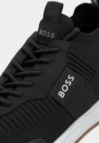 Baskets de sport noires avec une tige texturée, des lacets et un branding. Comprend une semelle blanche avec une semelle en caoutchouc et des détails galbés subtils.