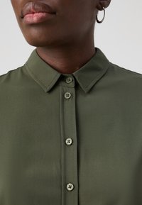 Olivenfarget skjorte med krage og knapper foran, med matchende knapper og en glatt tekstur. Nærbilde som fokuserer på halslinning og knapper.