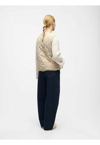 Beige gewatteerde vest met een staande kraag, gelaagd over een witte gestructureerde trui, gecombineerd met een losse donkerblauwe broek en lichte schoenen.