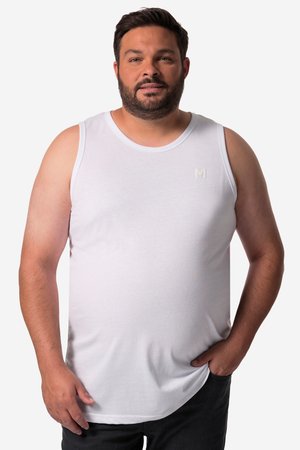 Man with beard wearing a white sleeveless tank top and black pants, standing with one hand in pocket against a plain white background.