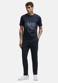 Dark blue t-shirt with graphic print, paired with dark jogger pants and white sneakers. Features short sleeves and a relaxed fit.