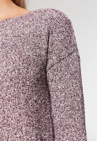 Pullovertröja i kastanjebrun och vit spräcklig stickning. Har en bred halsringning och långa ärmar med en avslappnad passform. Strukturerad tygdetalj synlig.