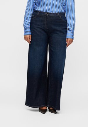 Femme portant un jean large bleu foncé, une chemise à manches longues rayée bleue et des chaussures pointues noires, debout sur fond blanc.
