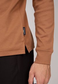 Chemise à manches longues marron avec un ourlet fendu sur le côté, texture douce et étiquette de marque visible sur le côté gauche près de la couture inférieure.