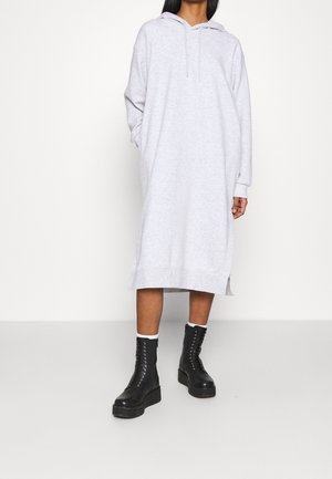 Osoba ubrana w jasnoszarą, długą sukienkę z kapturem, do której założono czarne buty na platformie z sznurowaniem oraz białe skarpetki do kostki.