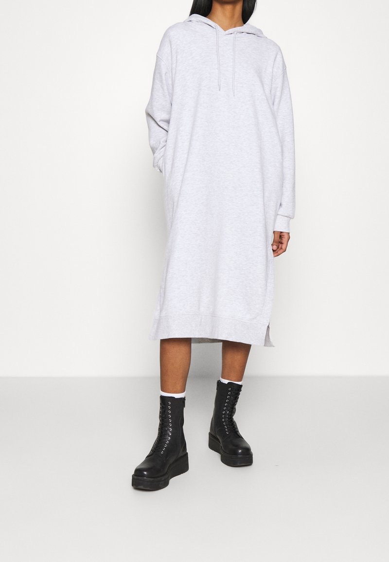 Personne portant une longue robe à capuche gris clair associée à des bottes à plateforme noires à lacets et des chaussettes blanches montant jusqu'aux chevilles.