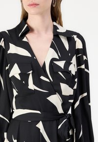 Black wrap dress with white abstract patterns, long sleeves, notch collar, two chest pockets, and a waist tie. Smooth, lightweight fabric.