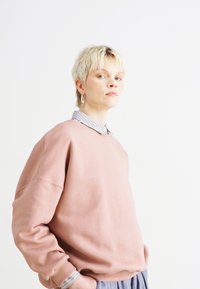 Felpa rosa oversize, dalla trama morbida, con scollatura rotonda e spalle abbassate, indossata sopra una camicia a righe con colletto, impreziosita da un orecchino ad anello in argento.