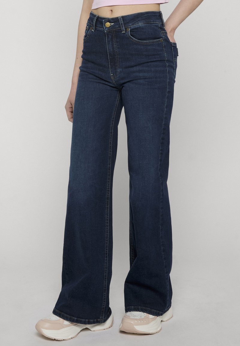 Femme portant un jean large taille haute bleu foncé avec un haut court rose et des baskets blanches, debout devant un fond uni.