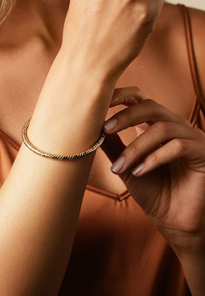 Vrouw draagt een goud- en zilverkleurige gedraaide armband aan de linkerpols, past deze aan met de rechterhand, gekleed in een bruin topje met spaghettibandjes.