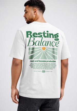 Man wearing white T-shirt with green text "Resting Balance" and a sunburst design promoting mental health and rest.