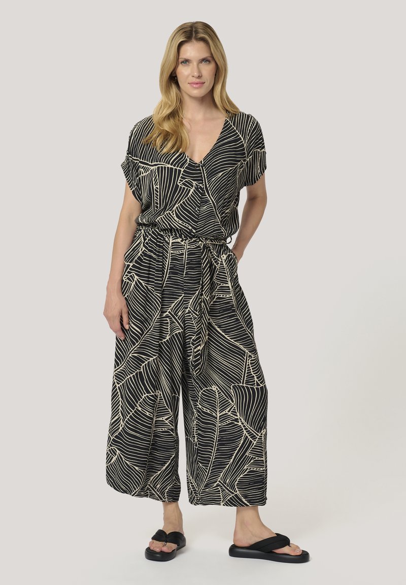 Svartvit mönstrad jumpsuit med V-ringning, korta ärmar, elastisk midja och vida ben; i kombination med svarta flip-flopsandaler.