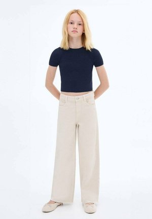 Navy geribbelde crop top, beige wijde broek en lichte schoenen; model staat met de armen langs het lichaam tegen een effen achtergrond.