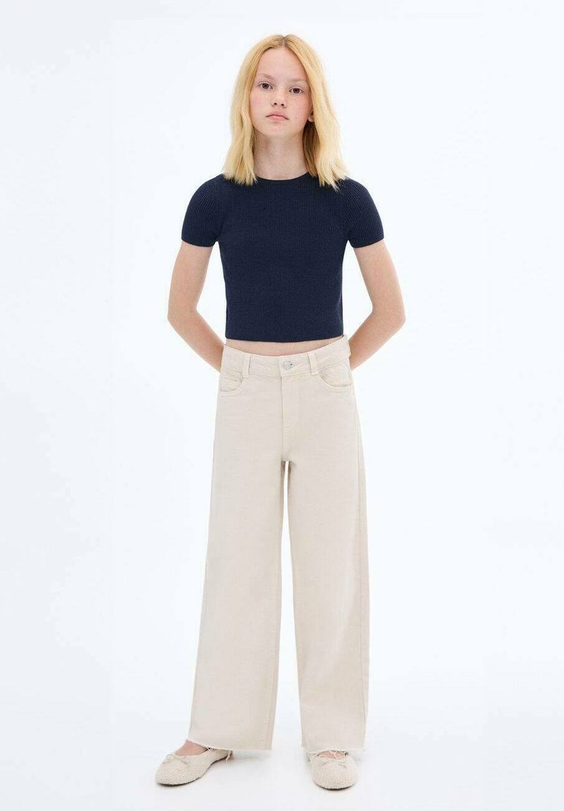 Top corto a righe color navy, pantaloni larghi beige e calzature chiare; il modello è in piedi con le braccia lungo i fianchi su uno sfondo semplice.