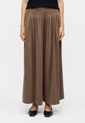 Vrouw draagt bruine wijde geplooide broek en zwarte platte schoenen, staand tegen een effen lichte achtergrond.