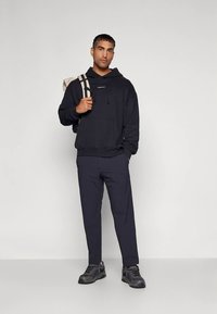 Άνδρας με μαύρη κουκούλα και σκούρα παντελόνια, κρατώντας τη θήκη ενός μπεζ σακιδίου με το ένα χέρι, στέκεται μπροστά σε απλή ανοιχτόχρωμη φόντο.