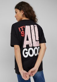 Zwart katoenen T-shirt met een grote, versleten grafische print in roze en wit op de achterkant met de tekst "IT'S ALL GOOD". Korte mouwen, losse pasvorm.