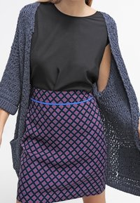 Stickad grå cardigan över en svart topp, matchad med en kjol med blå accenter som har ett geometriskt mönster i lila, röd och vit nyanser.