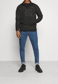 Veste noire zippée avec une poche en mesh et une taille élastique, associée à un jean bleu clair et des baskets noires.