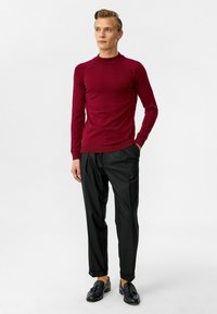 Pull en tricot bordeaux avec un col montant et des manches longues, associé à un pantalon noir plissé et des mocassins noirs. Texture lisse, coupe ajustée.