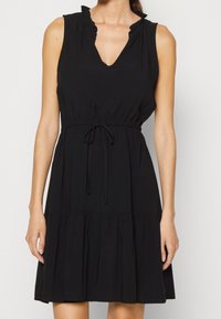 Svart ärmlös klänning med v-ringning, volangiga axlar, dragsko i midjan och en lager-på-lager kjol. Tillverkad av mjukt tyg med en slät yta.