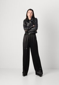Una donna sta in piedi con sicurezza, le braccia incrociate, indossando una giacca di pelle nera e pantaloni larghi neri, su uno sfondo chiaro e semplice.