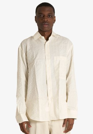 Giovane uomo che indossa una camicia oversize color crema con tessuto strutturato, con tasca frontale e pantaloni chiari abbinati, in piedi su sfondo bianco.